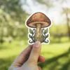 A whimsical mushroom sticker featuring a red-capped mushroom with intricate swirling patterns and green leaves at its base.