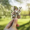 A whimsical garden gnome with a white beard, olive green hat, and beige jacket stands on wooden crates, holding onto grapevines.