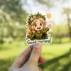 A cheerful blonde chibi elf girl wearing a flower crown and green tunic waves hello under a bright sun icon with 'Good morning!' text, vinyl sticker.