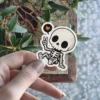 Cartoon skeleton making peace sign, speech bubble orange heart, black eye sockets, detailed bones, white border, vinyl sticker.