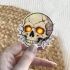 Cracked human skull, glowing yellow eyes, intricate spider web background, gothic horror vinyl sticker