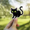 Cartoon black cat, piercing lime green eyes, arched tail, whimsical art style, vinyl sticker