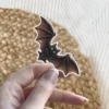 Small black bat, large golden eyes, spread wings, stylized illustration, vinyl sticker.