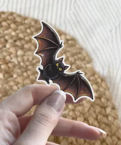 Small black bat, large golden eyes, spread wings, stylized illustration, vinyl sticker.