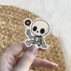 Chibi skeleton character, sitting, making peace sign gesture, black speech bubble, orange heart icon, black outlines, vinyl sticker.