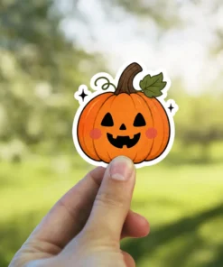 Orange pumpkin sticker, black carved eyes, smiling mouth, green stem leaf, pink blush cheeks, tiny sparkles vinyl sticker.