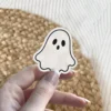 Adorable cartoon ghost sticker, white body, two black circular eyes, small oval black mouth, distinct black outline, vinyl sticker.