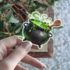 A playful frog peeks over the edge of a bubbling green cauldron, a wooden spoon with a symbol rests inside, detailed illustration, vinyl sticker.