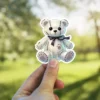 Ghostly white teddy bear, black stitched details, dark bow tie, gothic aesthetic, vinyl sticker