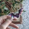 Cartoon purple bat, iridescent holographic wing details, sharp fangs, yellow eyes, gothic vinyl sticker.