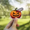 A cartoon jack-o-lantern with glowing yellow eyes and a jagged mouth is pierced by a bloody chainsaw, set against a blurred background, vinyl sticker.