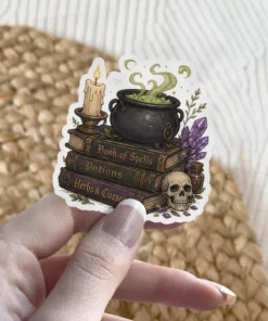 Stacked spell books, smoking cauldron, lit candle, purple crystals, human skull, potion bottle, green foliage vinyl sticker.