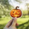 Orange jack-o'-lantern, evil glowing eyes, grinning mouth, cracked pumpkin skin, twisted brown stem, black outline, white border, vinyl sticker.