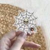 Skull centered on dew-covered spider web with small glowing orange spider, vinyl sticker.
