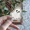 White ghost sticker, classic wavy shape, three black oval eyes, bold black outline, fun spooky vinyl sticker.