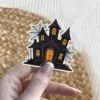 Dark silhouette haunted mansion, glowing orange windows, intricate spiderwebs, gothic architecture style, vinyl sticker