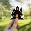Spooky haunted house silhouette, glowing orange windows, intricate black spiderwebs, artistic gothic vinyl sticker.