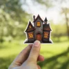 Haunted mansion illustration, boarded windows, glowing warm lights, dark gothic aesthetic, vinyl sticker